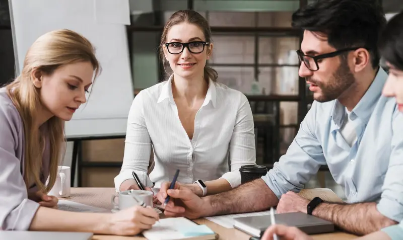 Four young professionals huddle at a desk: two writing notes, one consulting a laptop, and a smiling teammate in glasses looking toward the camera